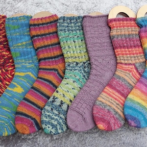 May include: A collection of seven colorful, hand-knitted socks. Each sock features a unique pattern and color scheme, including red, blue, purple, green, and rainbow hues. The socks are displayed on wooden forms, showcasing their intricate designs and textures.
