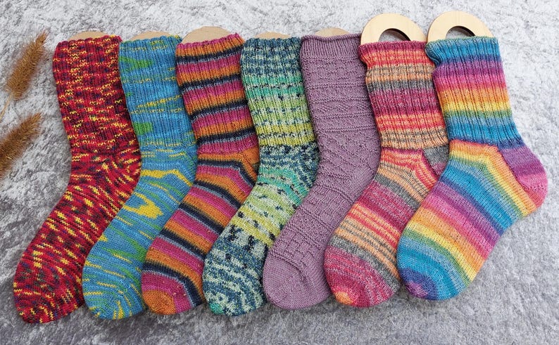 May include: A collection of seven colorful, hand-knitted socks. Each sock features a unique pattern and color scheme, including red, blue, purple, green, and rainbow hues. The socks are displayed on wooden forms, showcasing their intricate designs and textures.