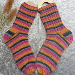 May include: A pair of colorful, striped socks. The socks feature horizontal bands of pink, orange, and navy blue. The socks are displayed on wooden sock forms. The background is a light gray, textured surface.