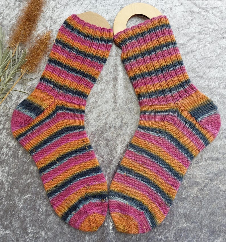 May include: A pair of colorful, striped socks. The socks feature horizontal bands of pink, orange, and navy blue. The socks are displayed on wooden sock forms. The background is a light gray, textured surface.