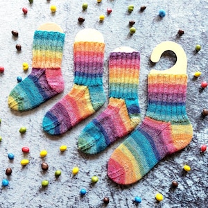Könnte beinhalten: Vier gestrickte Socken mit einem lebendigen Regenbogen-Farbverlaufsmuster mit horizontalen Streifen in Rot-, Orange-, Gelb-, Grün-, Blau- und Violetttönen. Jede Socke ist auf einem hellen Pappaufhänger präsentiert und in einer Reihe angeordnet.