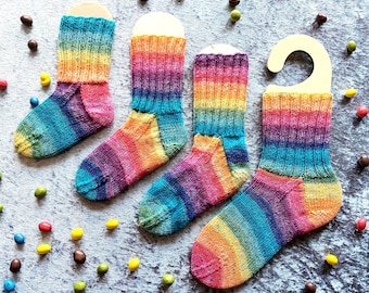 May include: Four knitted socks featuring a vibrant rainbow gradient pattern with horizontal stripes in shades of red, orange, yellow, green, blue, and purple. Each sock is displayed on a light-colored cardboard hanger, arranged in a row.