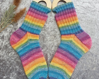 May include: A pair of knitted socks featuring a vibrant rainbow gradient pattern with horizontal stripes in shades of blue, green, yellow, orange, and pink. Each sock is displayed on a light-colored wooden sock blocker with a circular top opening.