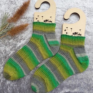 May include: A pair of hand-knitted socks with a green, yellow, and gray striped pattern. The socks are displayed on wooden hangers with a dandelion design. The socks are made of soft yarn and are perfect for keeping feet warm.
