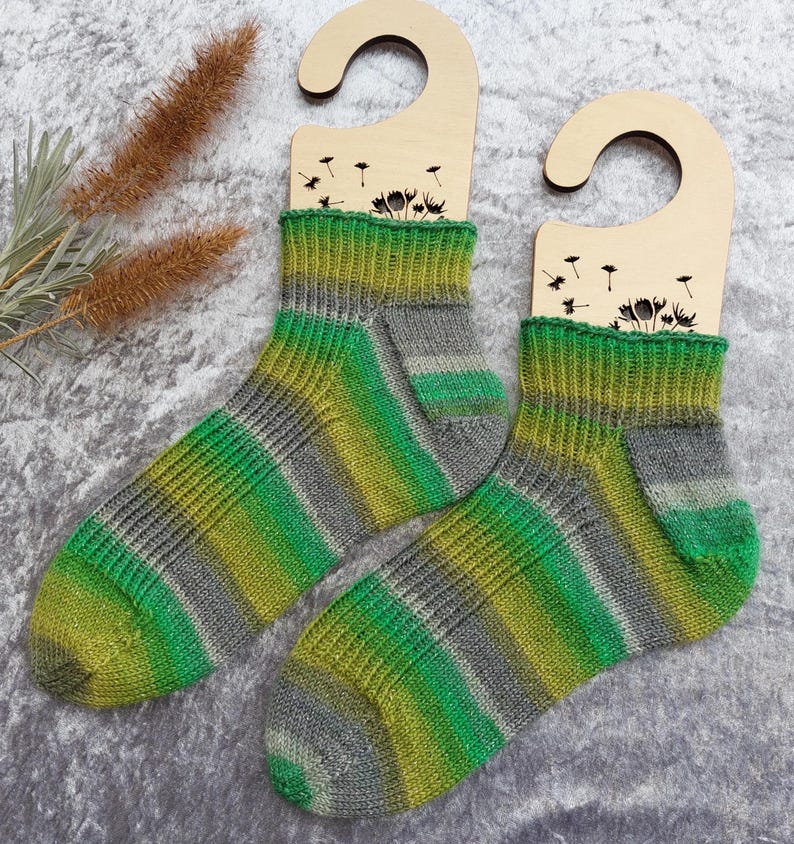 May include: A pair of hand-knitted socks with a green, yellow, and gray striped pattern. The socks are displayed on wooden hangers with a dandelion design. The socks are made of soft yarn and are perfect for keeping feet warm.