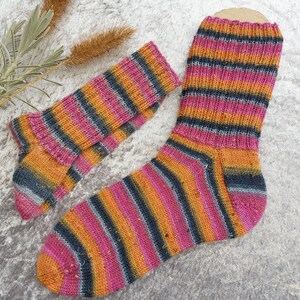 May include: A pair of hand-knitted socks with a vibrant striped pattern. The socks feature bands of pink, orange, and dark blue. The socks are made of a soft, textured yarn, perfect for keeping feet warm. The socks are a cozy accessory.