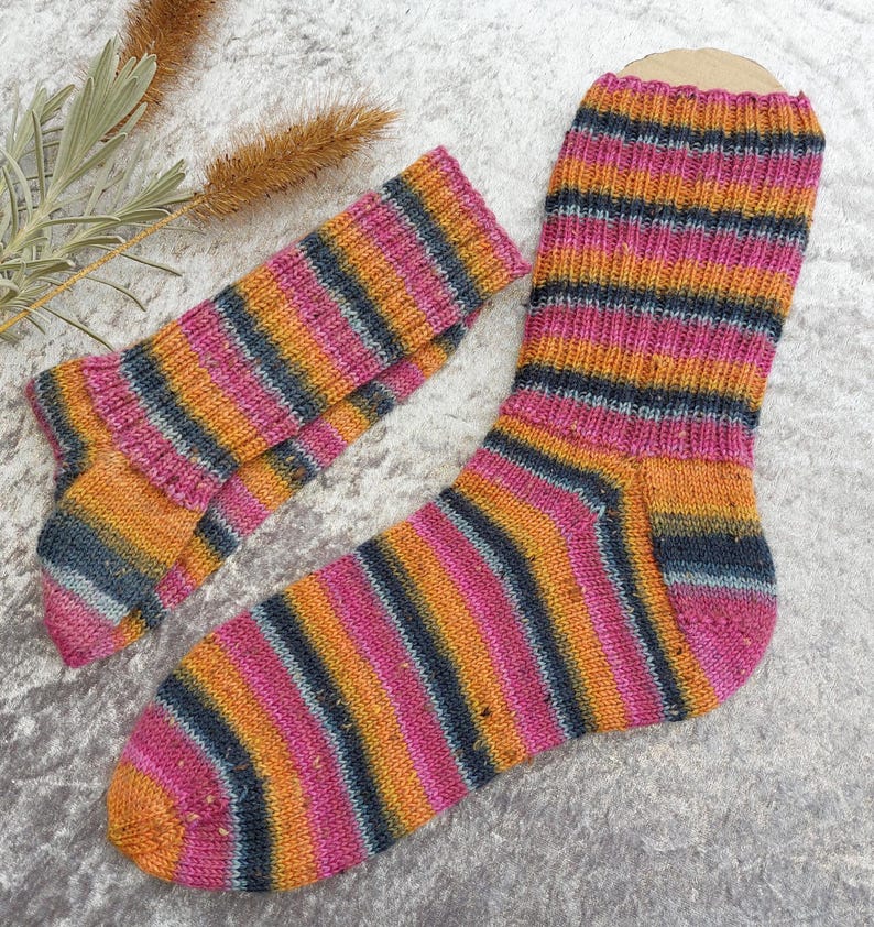 May include: A pair of hand-knitted socks with a vibrant striped pattern. The socks feature bands of pink, orange, and dark blue. The socks are made of a soft, textured yarn, perfect for keeping feet warm. The socks are a cozy accessory.