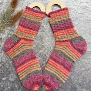 May include: A pair of colorful, striped socks with shades of pink, orange, and gray. The socks are displayed with wooden sock forms. The socks are made of a soft, knitted material.