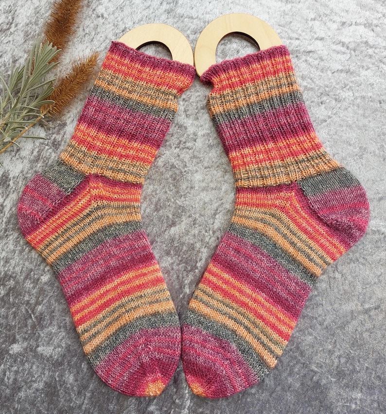 May include: A pair of colorful, striped socks with shades of pink, orange, and gray. The socks are displayed with wooden sock forms. The socks are made of a soft, knitted material.