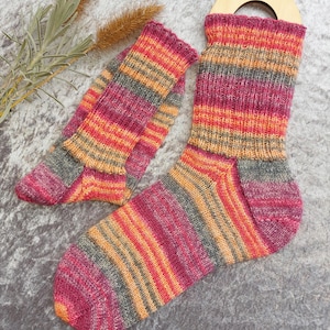 May include: A pair of colorful knitted socks with a matching cuff. The socks feature horizontal stripes in shades of pink, orange, gray, and yellow. The socks are displayed on a gray surface.