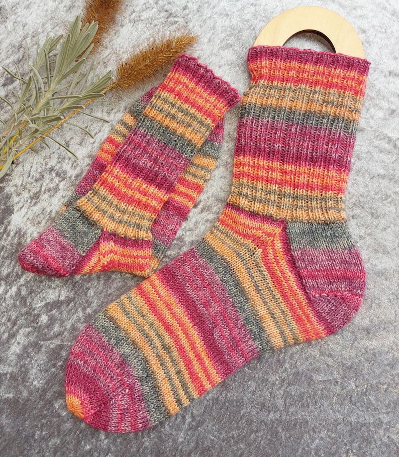 May include: A pair of colorful knitted socks with a matching cuff. The socks feature horizontal stripes in shades of pink, orange, gray, and yellow. The socks are displayed on a gray surface.