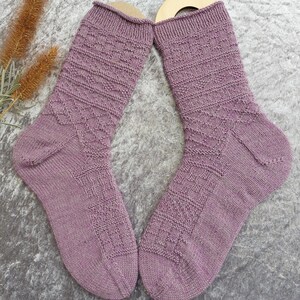May include: A pair of lavender-colored knitted socks with textured patterns. The socks feature a ribbed cuff, geometric designs on the leg, and a reinforced heel and toe. The socks are displayed on a gray surface.