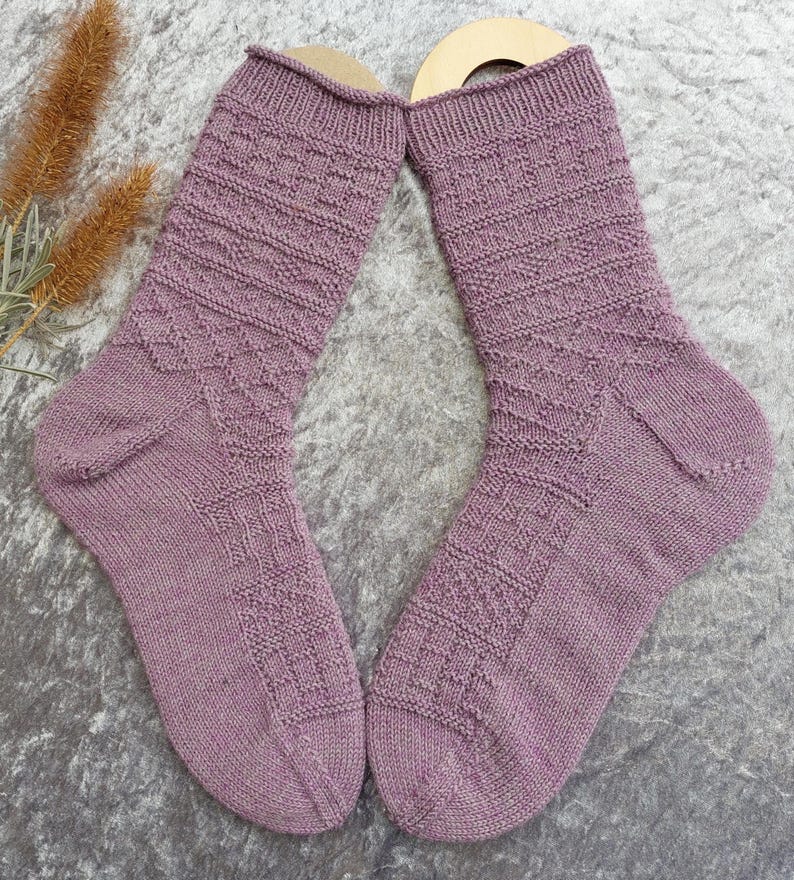 May include: A pair of lavender-colored knitted socks with textured patterns. The socks feature a ribbed cuff, geometric designs on the leg, and a reinforced heel and toe. The socks are displayed on a gray surface.