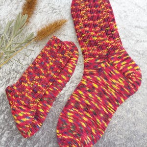 May include: A pair of knitted socks in a vibrant mix of red, yellow, blue, and green. The socks are made of a textured yarn and appear to be hand-knitted. The socks are displayed on a gray surface.