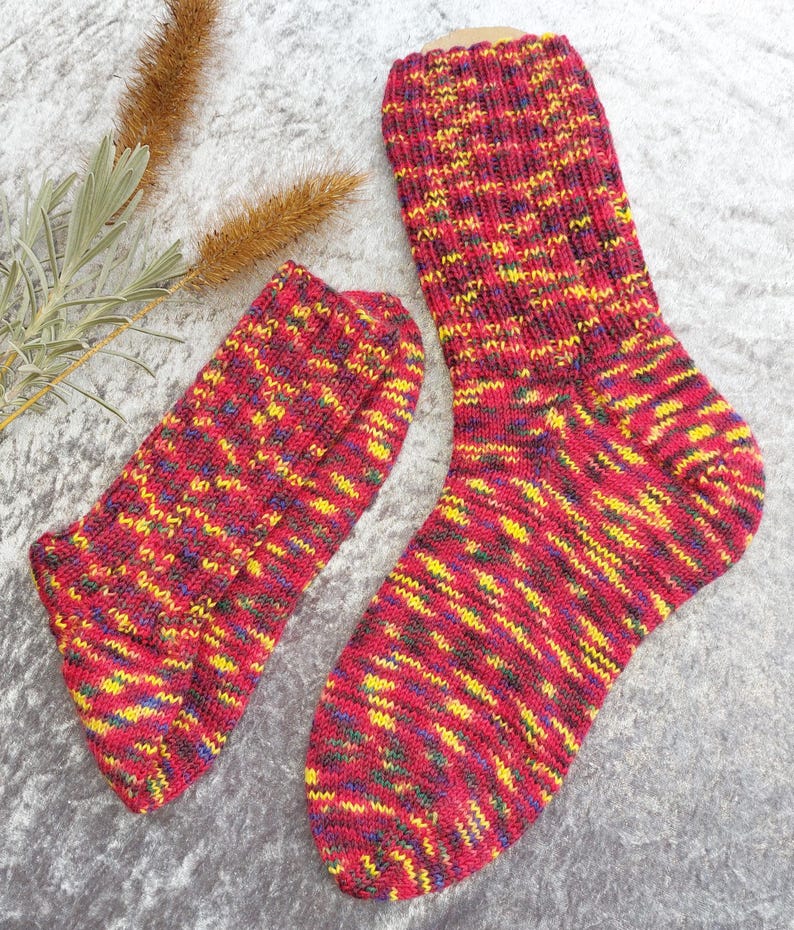 May include: A pair of knitted socks in a vibrant mix of red, yellow, blue, and green. The socks are made of a textured yarn and appear to be hand-knitted. The socks are displayed on a gray surface.