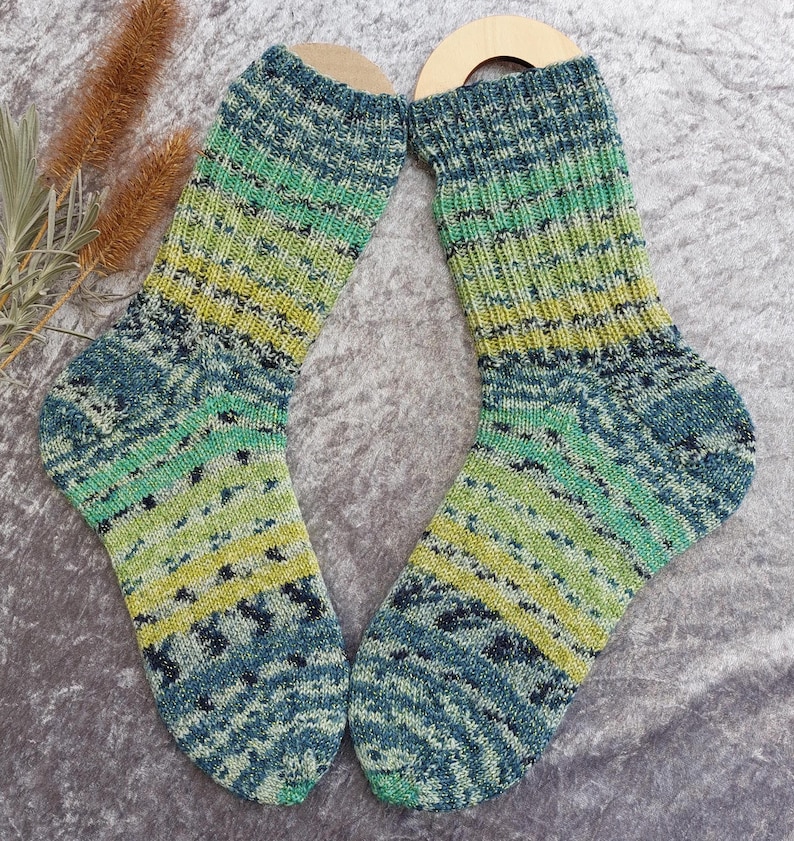 May include: A pair of hand-knitted socks in shades of blue, green, and yellow. The socks feature a ribbed design on the upper portion and a patterned design on the foot. The socks are displayed on wooden sock forms.