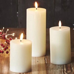 May include: Three white pillar candles with a flickering flame effect. The candles are different sizes, with the largest candle at the back and the smallest candle at the front.