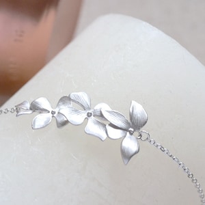 Pode incluir: Um delicado bracelete de prata com um grupo de cinco pingentes de flores estilizadas conectados por uma corrente de prata.