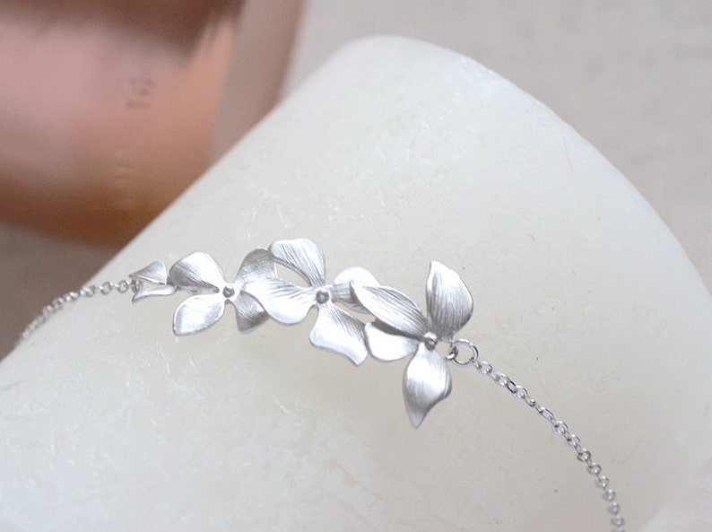 Pode incluir: Um delicado bracelete de prata com um grupo de cinco pingentes de flores estilizadas conectados por uma corrente de prata.