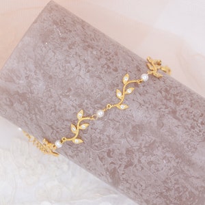 Peut inclure: Un collier délicat en or avec un motif de feuilles et de petites perles blanches. Le collier est posé sur une surface texturée de couleur brun clair.