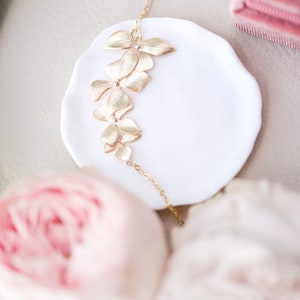 Pode incluir: Um colar de ouro com uma corrente delicada e um design floral. O colar &eacute; apresentado em um prato branco com uma flor rosa no fundo.
