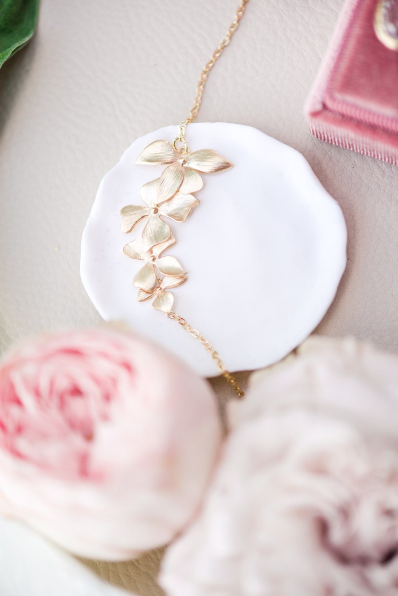 Pode incluir: Um colar de ouro com uma corrente delicada e um design floral. O colar &eacute; apresentado em um prato branco com uma flor rosa no fundo.