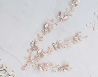 May include: A rose gold bridal hair vine with leaf and flower details. It features pale pink fabric leaves, clear crystals, and small pearl beads. The vine is arranged on a flexible wire, suitable for various hairstyles. The design is elegant and refined.