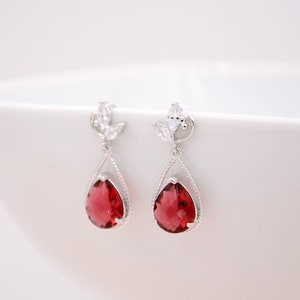May include: A pair of silver dangle earrings with a teardrop-shaped red gemstone and a small cluster of clear cubic zirconia stones.