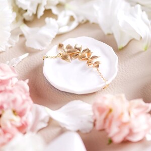 Pode incluir: Um colar de ouro com um delicado design floral, descansando sobre um prato de cer&acirc;mica branca. O colar est&aacute; rodeado de p&eacute;talos de flores brancas e rosadas.