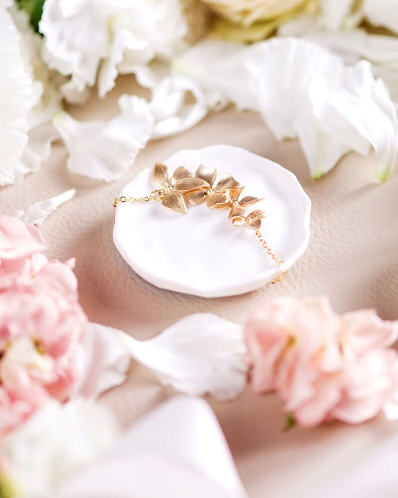 Pode incluir: Um colar de ouro com um delicado design floral, descansando sobre um prato de cer&acirc;mica branca. O colar est&aacute; rodeado de p&eacute;talos de flores brancas e rosadas.