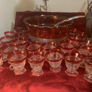 Puede incluir: Un juego de 18 vasos de ponche vintage de vidrio con borde rojo y base transparente. Los vasos están dispuestos en círculo alrededor de un gran tazón de ponche de vidrio rojo con base transparente.
