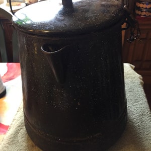 Puede incluir: Cafetera de camping de esmalte negro con tapa y asa de alambre. La cafetera tiene un acabado moteado.