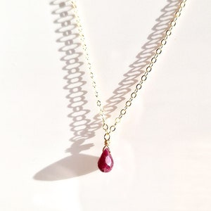 May include: A fine gold chain necklace featuring a teardrop-shaped, faceted ruby pendant. The ruby is a deep red hue and is wire-wrapped to the chain. The necklace is shown against a white backdrop, with shadows.