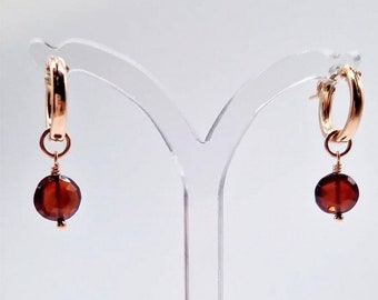 May include: A pair of rose gold hoop earrings with a faceted, round, dark red gemstone dangling from each hoop. The earrings are displayed on a clear stand against a white background. The gemstones appear to be approximately 0.5 inches in diameter.
