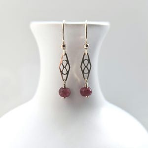 May include: A pair of gold-coloured earrings. Each earring features a geometric design and a faceted, round, red gemstone. The earrings are displayed on a white surface. The design is delicate and elegant.