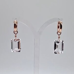 May include: A pair of rose gold hoop earrings with clear, rectangular, faceted crystal pendants. The earrings are displayed on a clear stand against a white background. The crystals are suspended from the hoops by delicate wire.