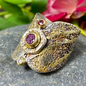 May include: Artisan-crafted ring with a textured silver and gold design. The ring features three gemstones: a large central ruby, a smaller ruby, and a small diamond. The ring's unique design showcases a blend of textures and colours.