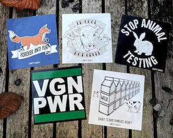 May include: Five square stickers with black and white illustrations and text promoting veganism and animal rights. The stickers feature a fox, a cow, a pig, a rabbit, and a cow drinking milk. The text on the stickers includes "Forever Anti Fur", "Be Cool Not Cruel", "Stop Animal Testing", "VGN PWR", and "Dairy Tears Families Apart".