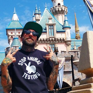 Peut inclure: Une personne porte un t-shirt bleu marine avec le texte "STOP ANIMAL TESTING" et un graphique de lapin blanc. La personne porte des lunettes de soleil et une casquette noire à visière verte. En arrière-plan, un château aux toits bleus.
