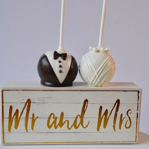 May include: Two cake pops decorated as a bride and groom, on a white wooden box with the words "Mr and Mrs" in gold lettering.