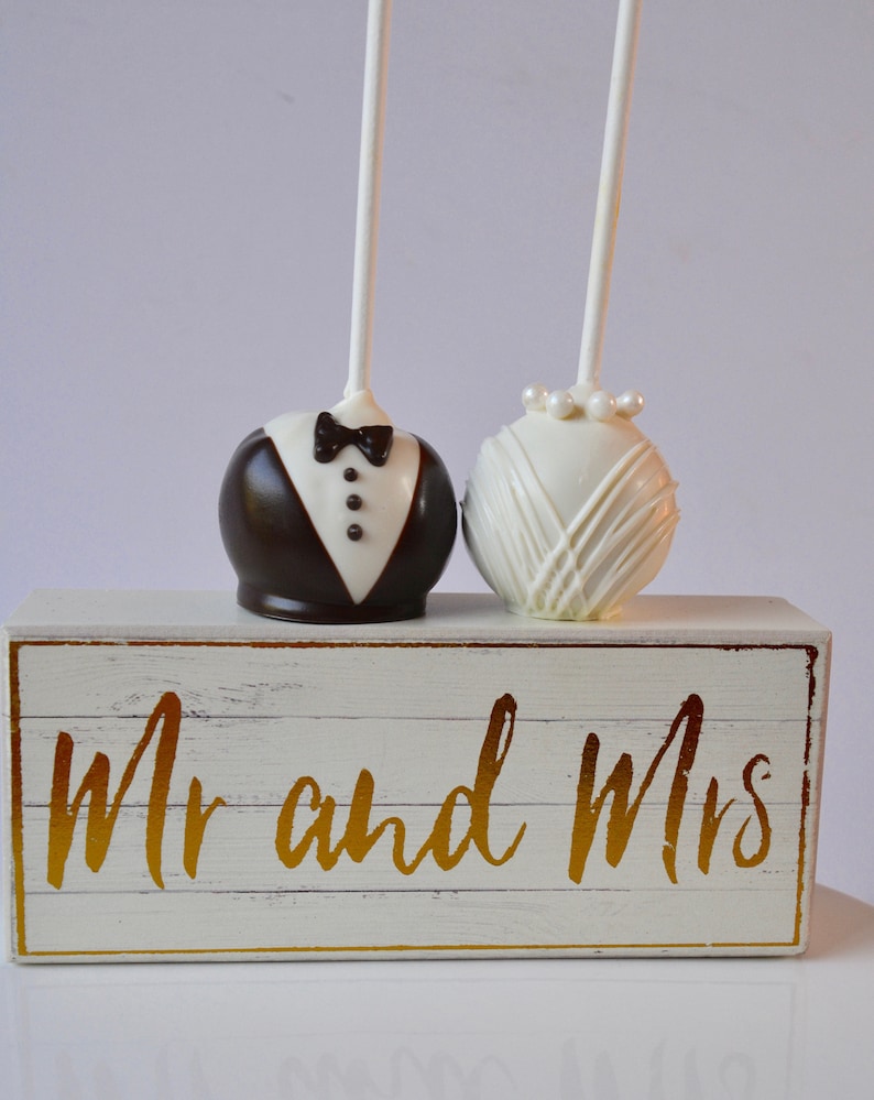 May include: Two cake pops decorated as a bride and groom, on a white wooden box with the words "Mr and Mrs" in gold lettering.
