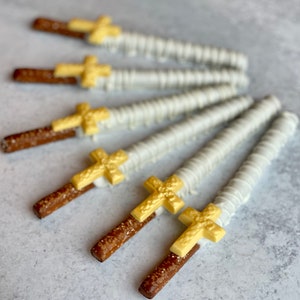 May include: A group of six pretzel rods dipped in white chocolate and topped with gold cross shapes. The pretzels are arranged in a fan shape on a gray surface.