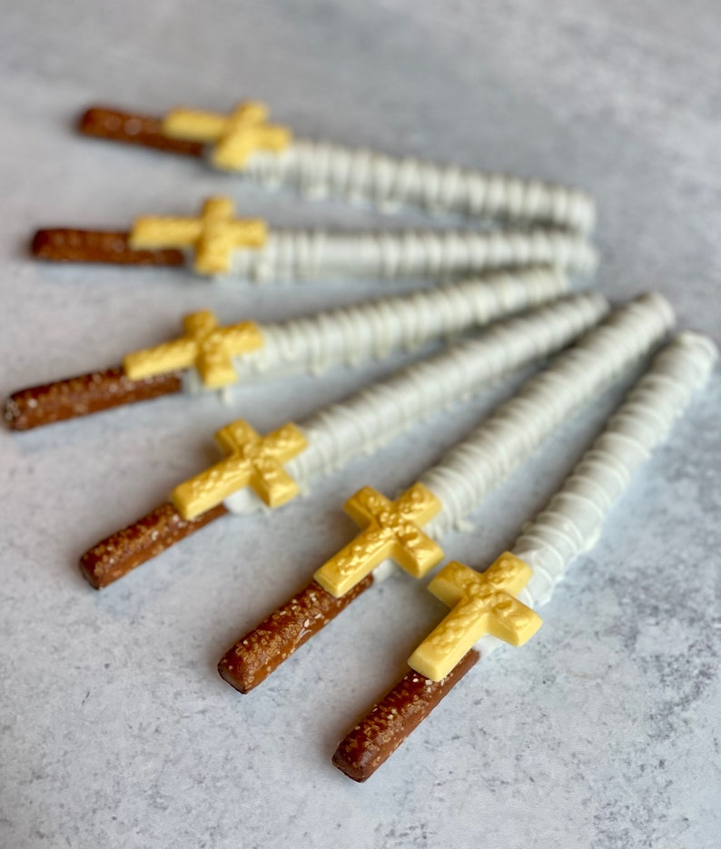 May include: A group of six pretzel rods dipped in white chocolate and topped with gold cross shapes. The pretzels are arranged in a fan shape on a gray surface.