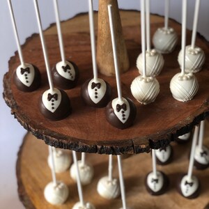 May include: A two-tiered wooden cake stand with white and brown cake pops. The cake pops are decorated with a tuxedo and a pearl design.