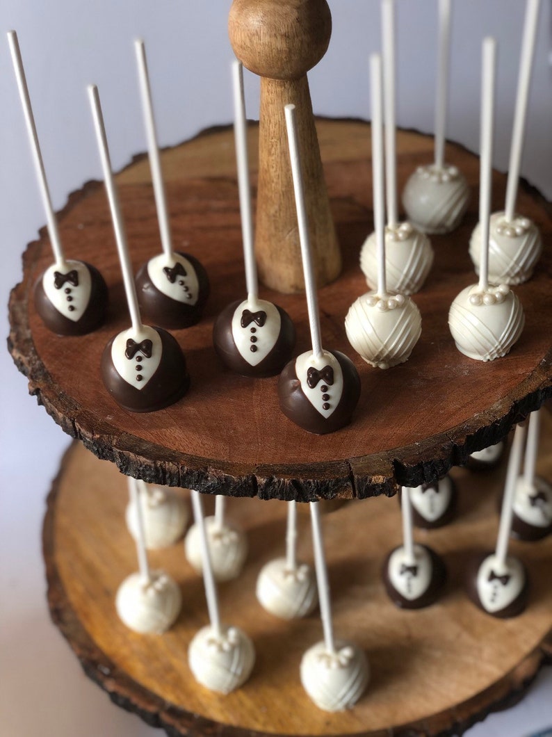 May include: A two-tiered wooden cake stand with white and brown cake pops. The cake pops are decorated with a tuxedo and a pearl design.