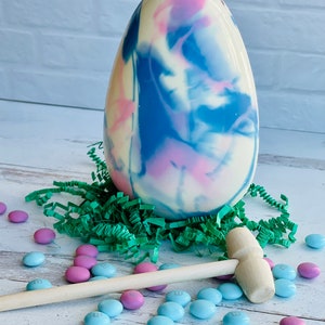 May include: A large white chocolate Easter egg with blue and pink marbling, sitting on green shredded paper. A wooden mallet and blue and pink M&Ms are scattered around the egg.