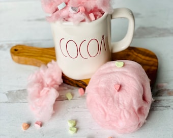 Puede incluir: Una taza de cerámica blanca con la palabra "COCOA" impresa en rojo en la parte delantera, llena de algodón de azúcar rosa y caramelos de colores en forma de corazón.