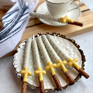 May include: Six white chocolate covered pretzel rods with gold cross toppers on a silver platter. The pretzels are arranged in a fan shape.