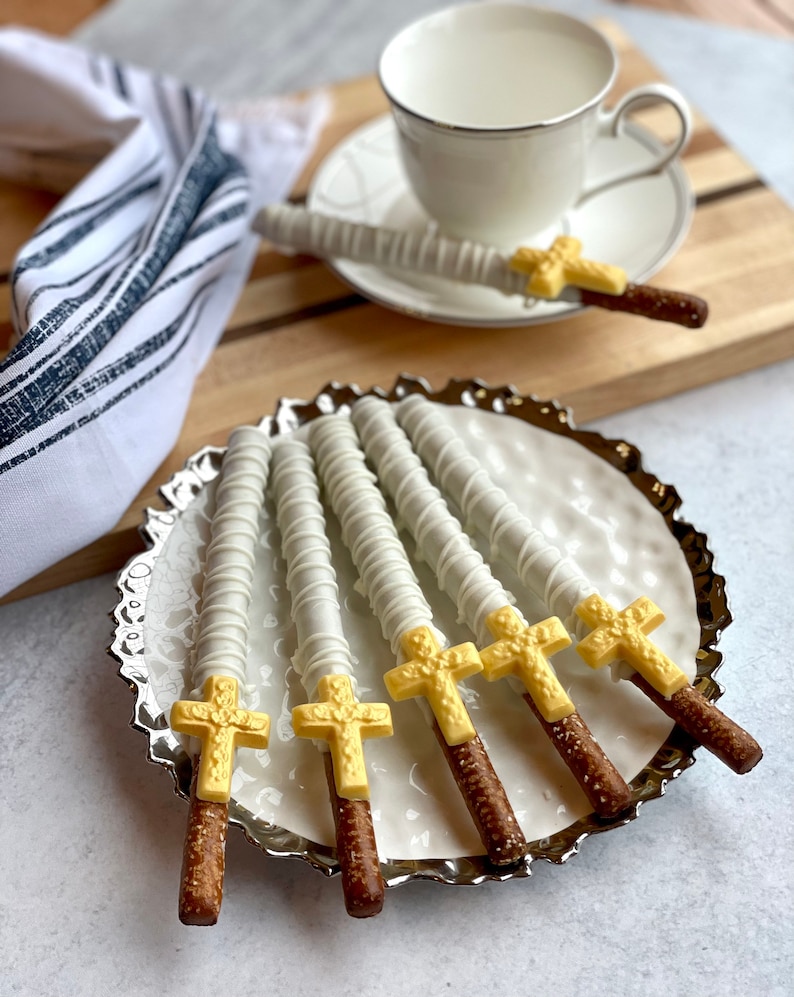 May include: Six white chocolate covered pretzel rods with gold cross toppers on a silver platter. The pretzels are arranged in a fan shape.
