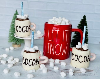 Puede incluir: Tres tazas de chocolate caliente de chocolate blanco con malvaviscos y pajitas azules, una taza roja con malvaviscos blancos que dice "Let It Snow", y malvaviscos blancos esparcidos sobre una superficie de madera blanca.